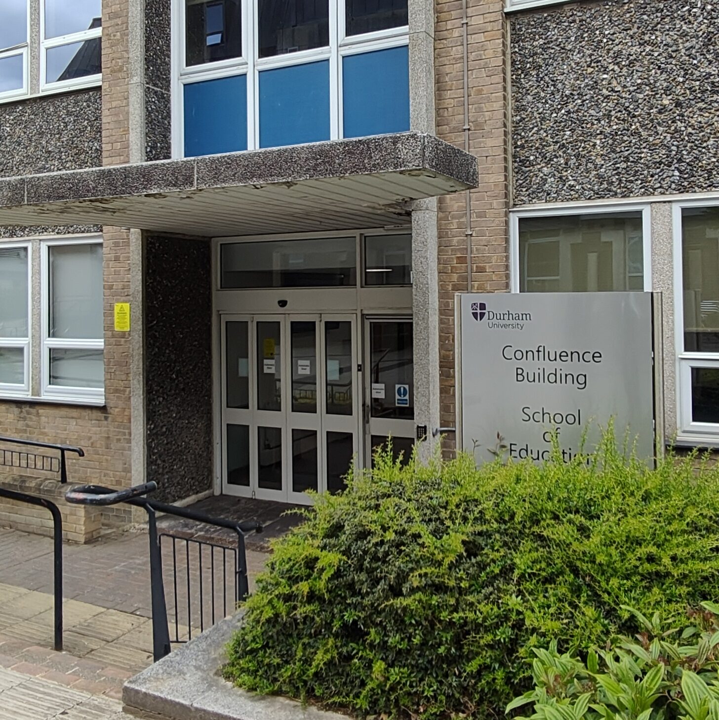 Entrance to Confluence Building. A set of double doors under a concrete awnings with a silver sign to the right which reads "Confluence Building: School of Education".