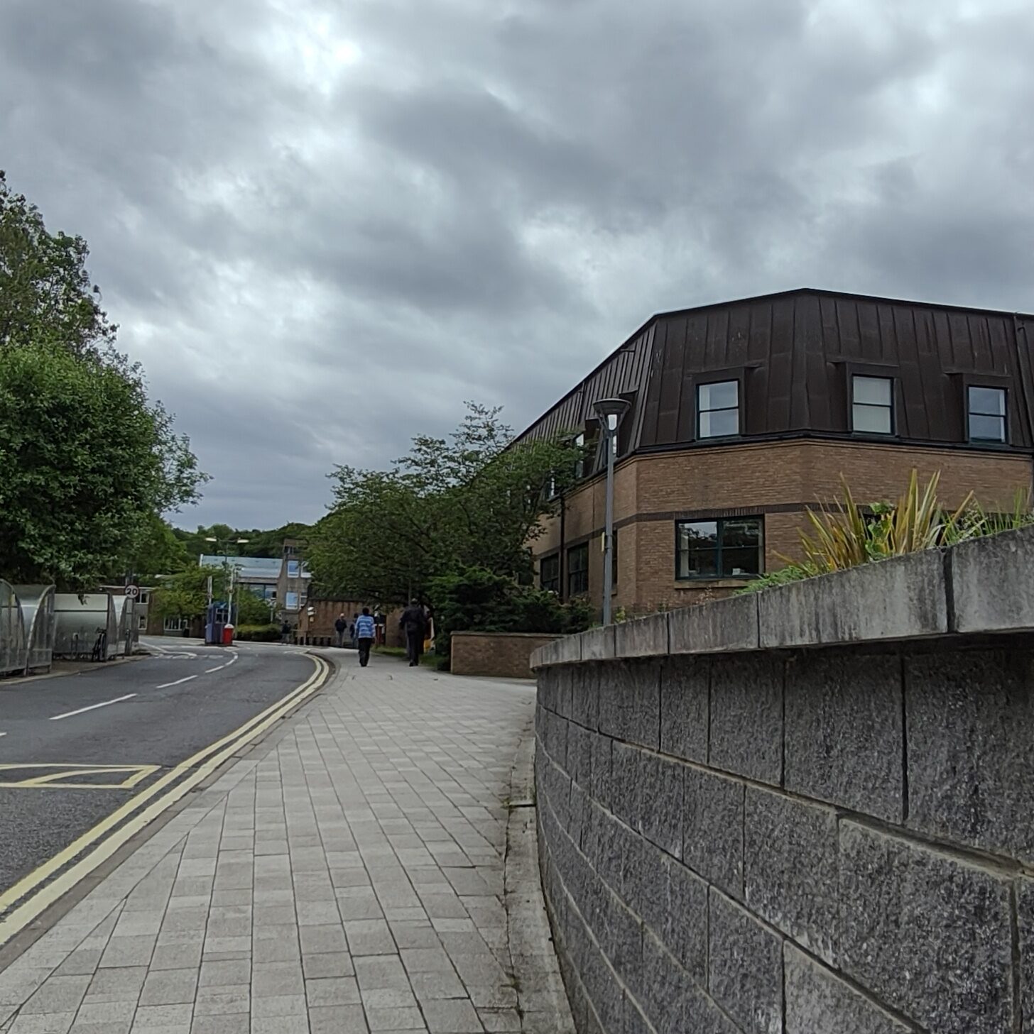 Stone-tiled path heading towards an red-bricked building with a brown roof. On the left-hand side is a road with double yellow lines.